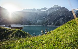 Two hikers look down to the Hochgebirgsstausee Kaprun and in the background you can see a hut and the snow-covered mountains