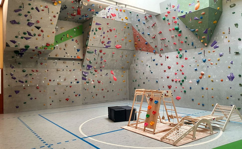 Indoor-Kletterhalle mit hohen Boulderwänden, bunten Griffen und Spielgeräten auf markiertem Hallenboden.
