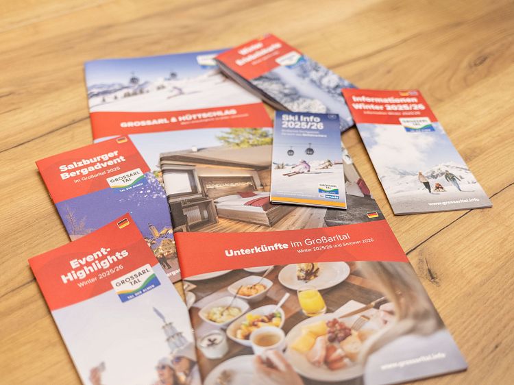 Mehrere bunte Broschüren zu Großarltal-Urlaub auf Holztisch zeigen Aktivitäten und Unterkünfte.