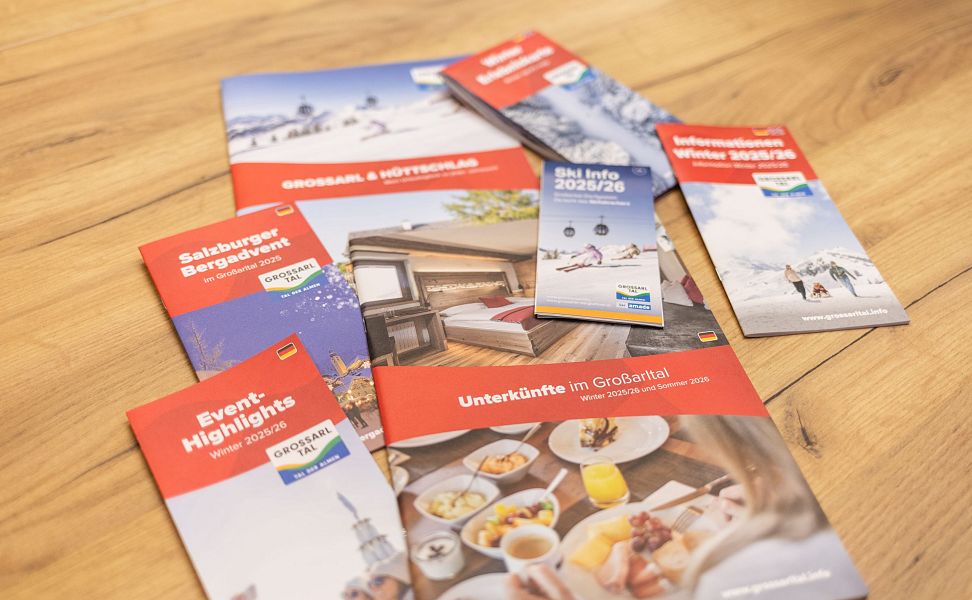 Mehrere bunte Broschüren zu Großarltal-Urlaub auf Holztisch zeigen Aktivitäten und Unterkünfte.