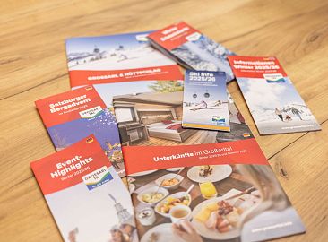 Mehrere bunte Broschüren zu Großarltal-Urlaub auf Holztisch zeigen Aktivitäten und Unterkünfte.