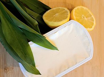 Bärlauch, ein Mörser, eine Zitronenhälfte und eine Schüssel mit Frischkäse stehen auf der Arbeitsplatte.